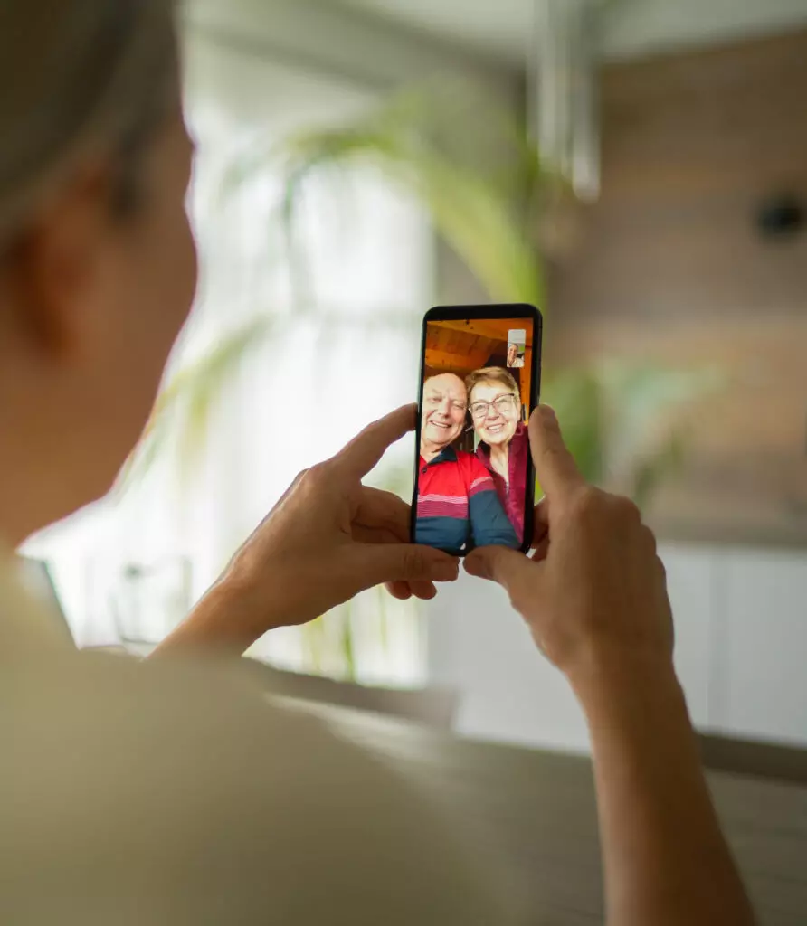 over the shoulder view mature adult woman talking with her smiling senior parents via mobile phone video telephony during corona quarantine in living room of house in times of social distancing to stay safe and healthy